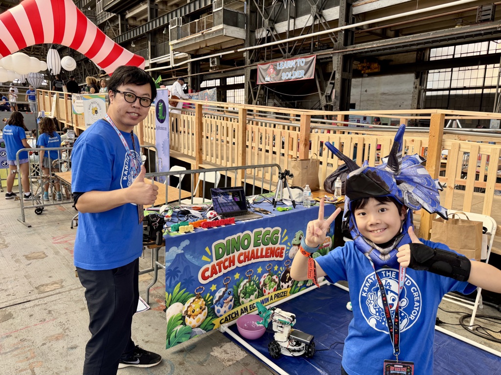 Kanata in a dinosaur hood and his dad giving a thumbs up in front of the DINO EGG Catch Challenge booth