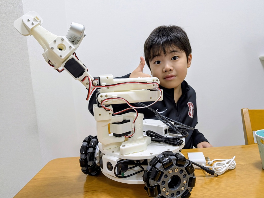 Kanata giving a thumbs up next to the LeKiwi robot and SO-ARM101 arm