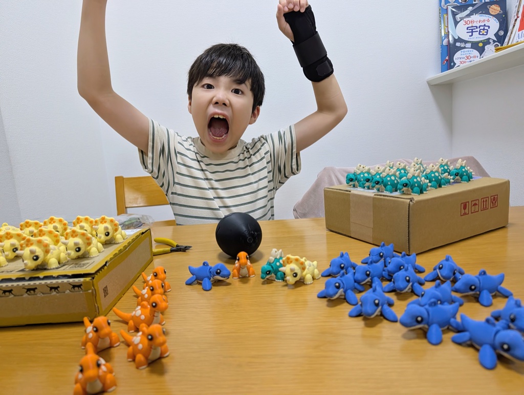Kanata cheering behind boxes of 3D-printed dinosaur keychains in yellow, orange, blue and teal
